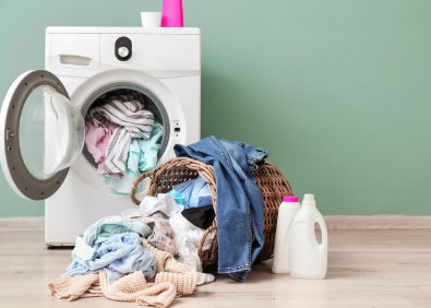 Offene Waschmaschine mit Wäschekorb, Kleidung und Waschmittel als Teaserbild zum Thema Wäsche richtig waschen und Textilpflege im Haushalt