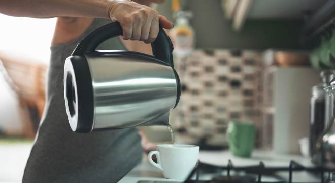 Person gießt heißes Wasser aus einem Wasserkocher in eine Tasse in der Küche als Teaserbild zum Thema Wasserkocher entkalken und reinigen