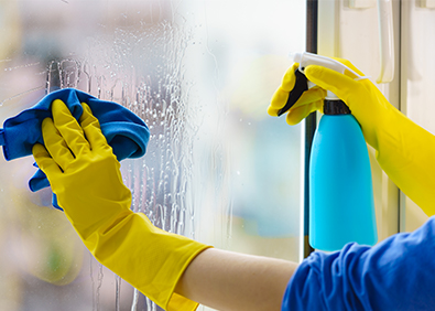 Person mit gelben Reinigungshandschuhen reinigt ein Fenster mit blauem Tuch und Sprühflasche für streifenfreie Glasreinigung