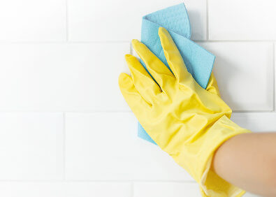 Hand mit gelbem Reinigungshandschuh wischt mit blauem Reinigungstuch über weiße Fliesen und Fugen zur hygienischen Reinigung im Bad oder in der Küche